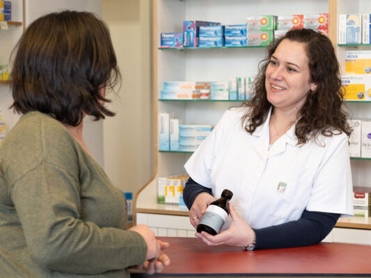 Apothekerin in weißem Kittel berät eine Kundin und hält eine Flasche mit Naturheilmittel.