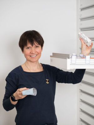 Apothekerin in dunkler Kleidung lächelt, während sie eine Medikamentenschublade öffnet.