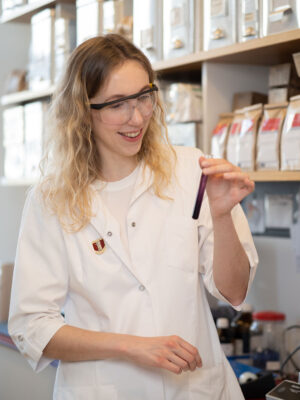 Junge Apothekerin mit Schutzbrille hält ein Reagenzglas mit einer dunklen Flüssigkeit.