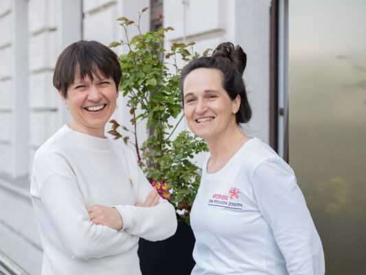 Zwei lachende Frauen vor der Apotheke, eine trägt ein Shirt mit dem Logo der Apotheke.